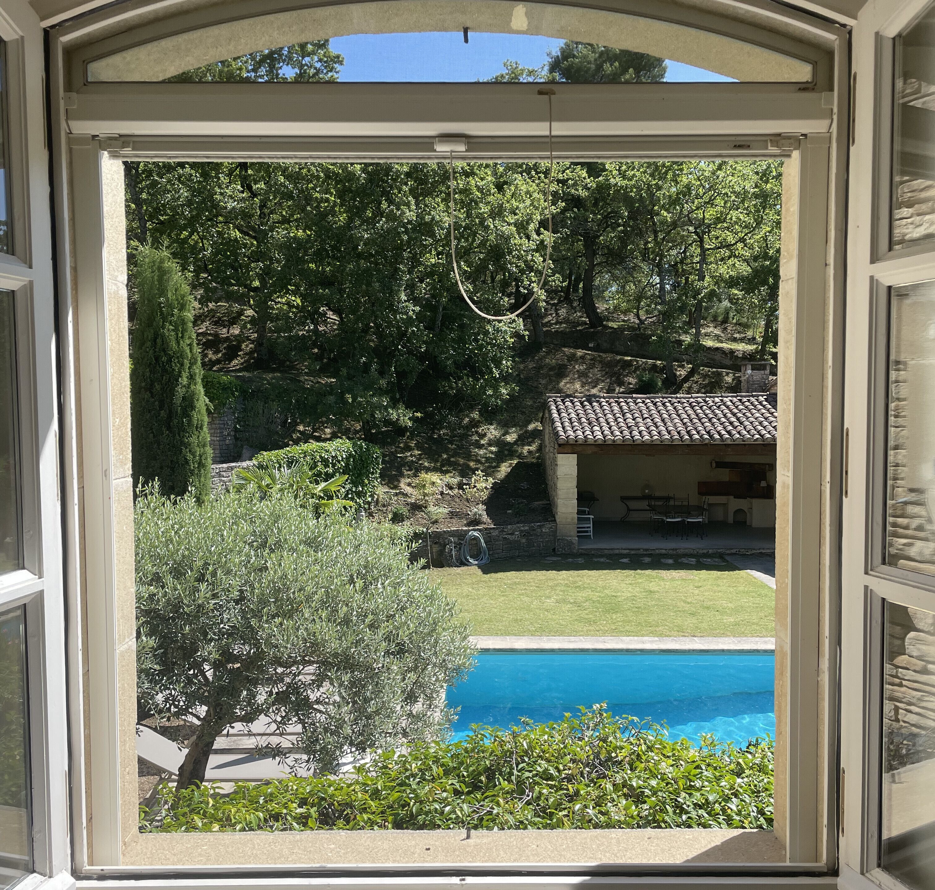 Une vue à travers une fenêtre sur une piscine, entourée de verdure et d’un bâtiment en pierre avec un toit en tuiles.