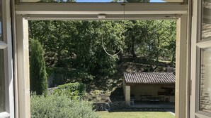 A view through a window of a swimming pool, surrounded by greenery and a stone building with a tiled roof. - Propriété d'exception au coeur du Luberon (Robion)