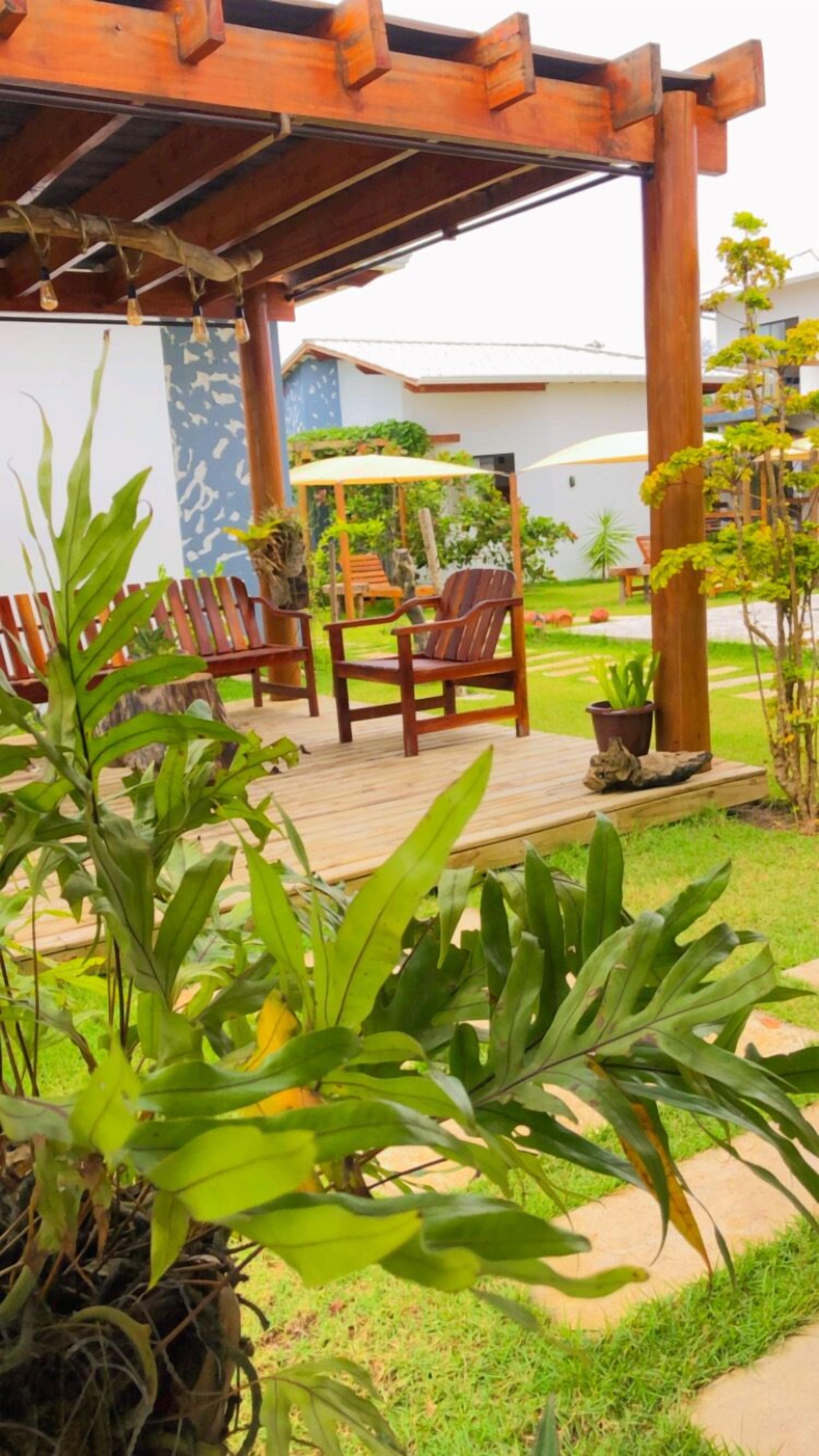 Un área de estar al aire libre con techo, con sillas y mesa de madera, rodeada de abundante follaje y plantas en macetas.