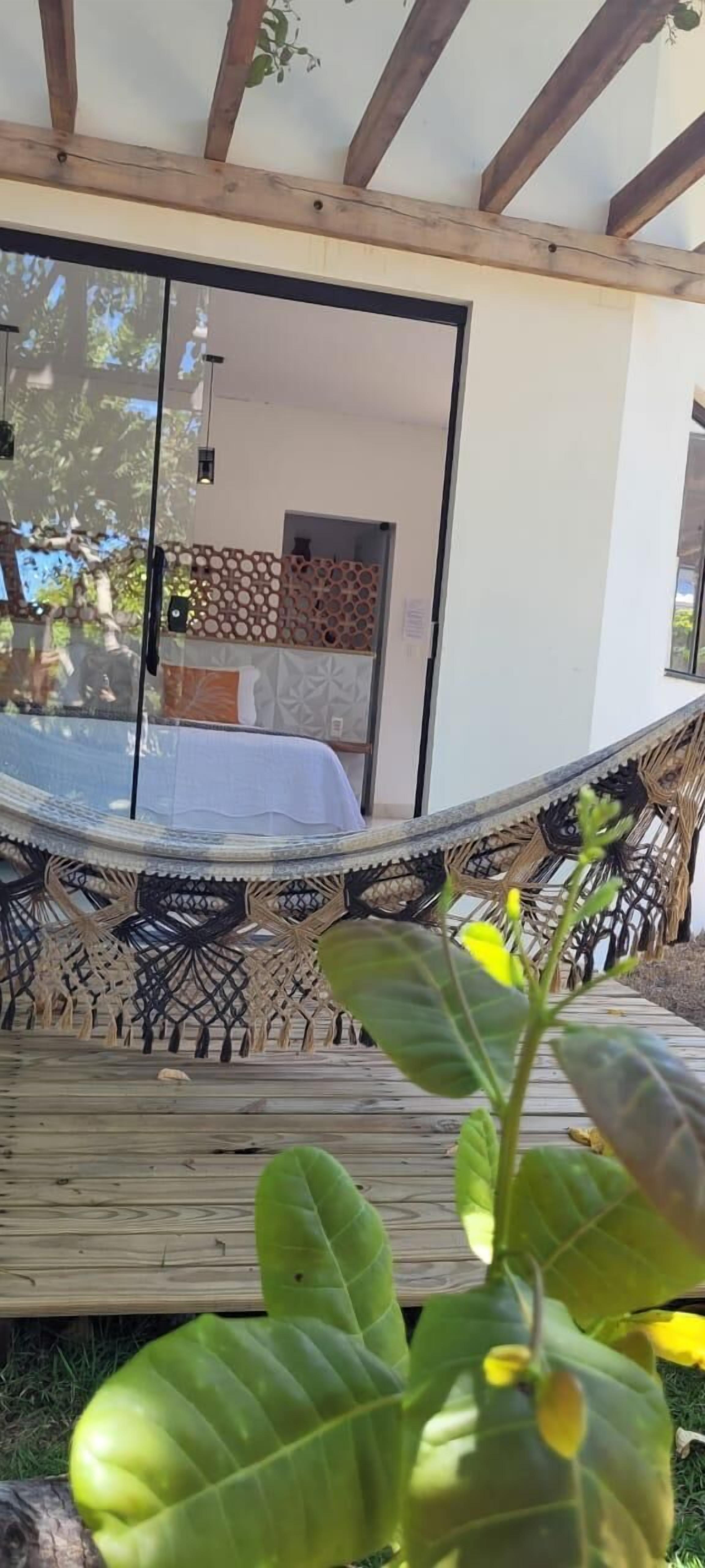 Un fauteuil suspendu sous une structure en poutres de bois, une porte vitrée donnant sur une pièce aux murs décorés, et une terrasse en bois avec de la verdure au premier plan.