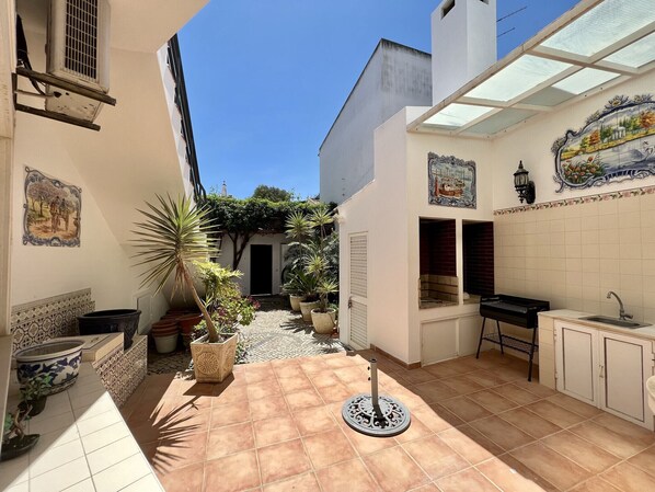 A sunlit courtyard with a tiled floor, potted plants, a decorative fountain, and a view of the street.