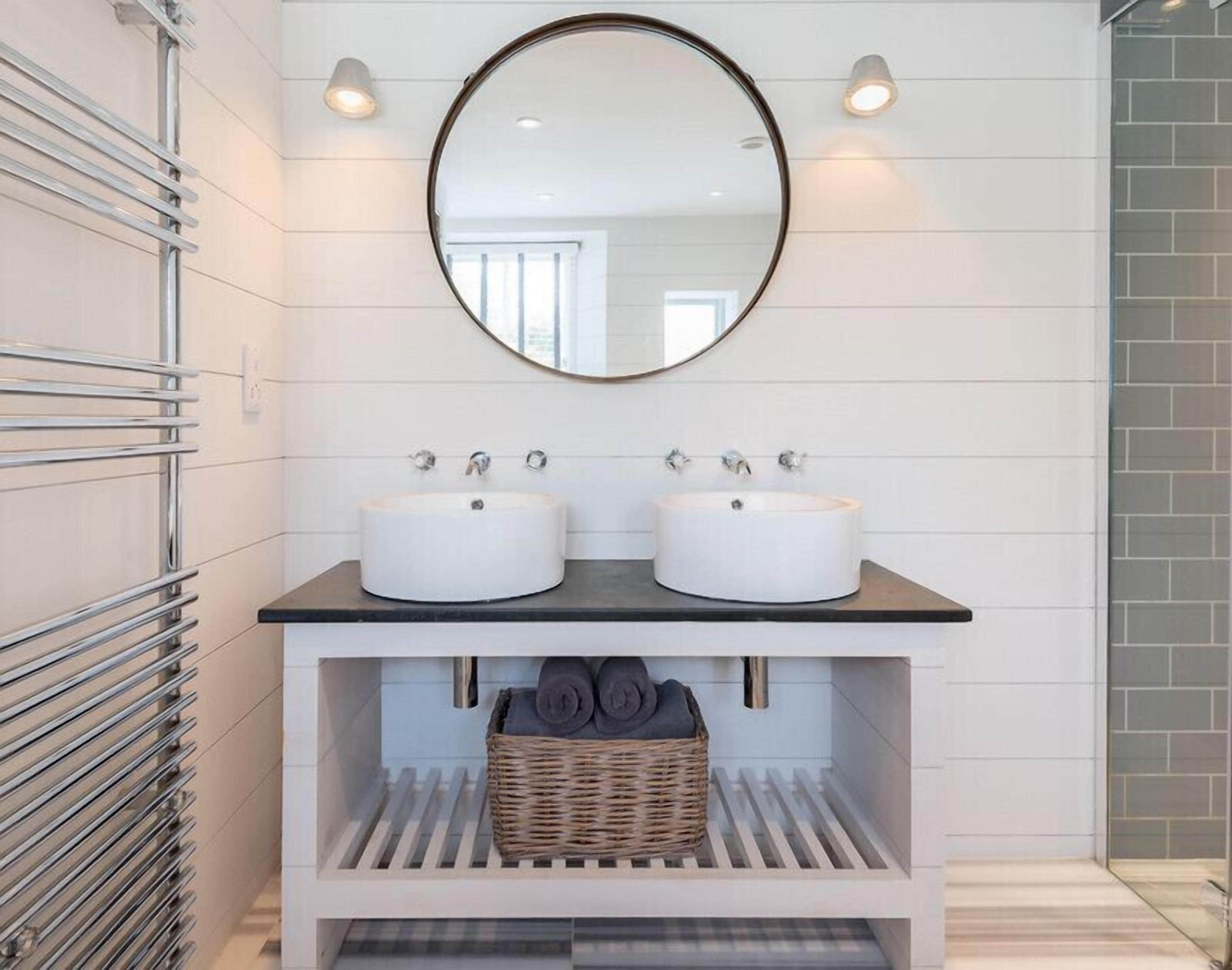 A bathroom with two white sinks, a round mirror, and a wicker basket under the sink.