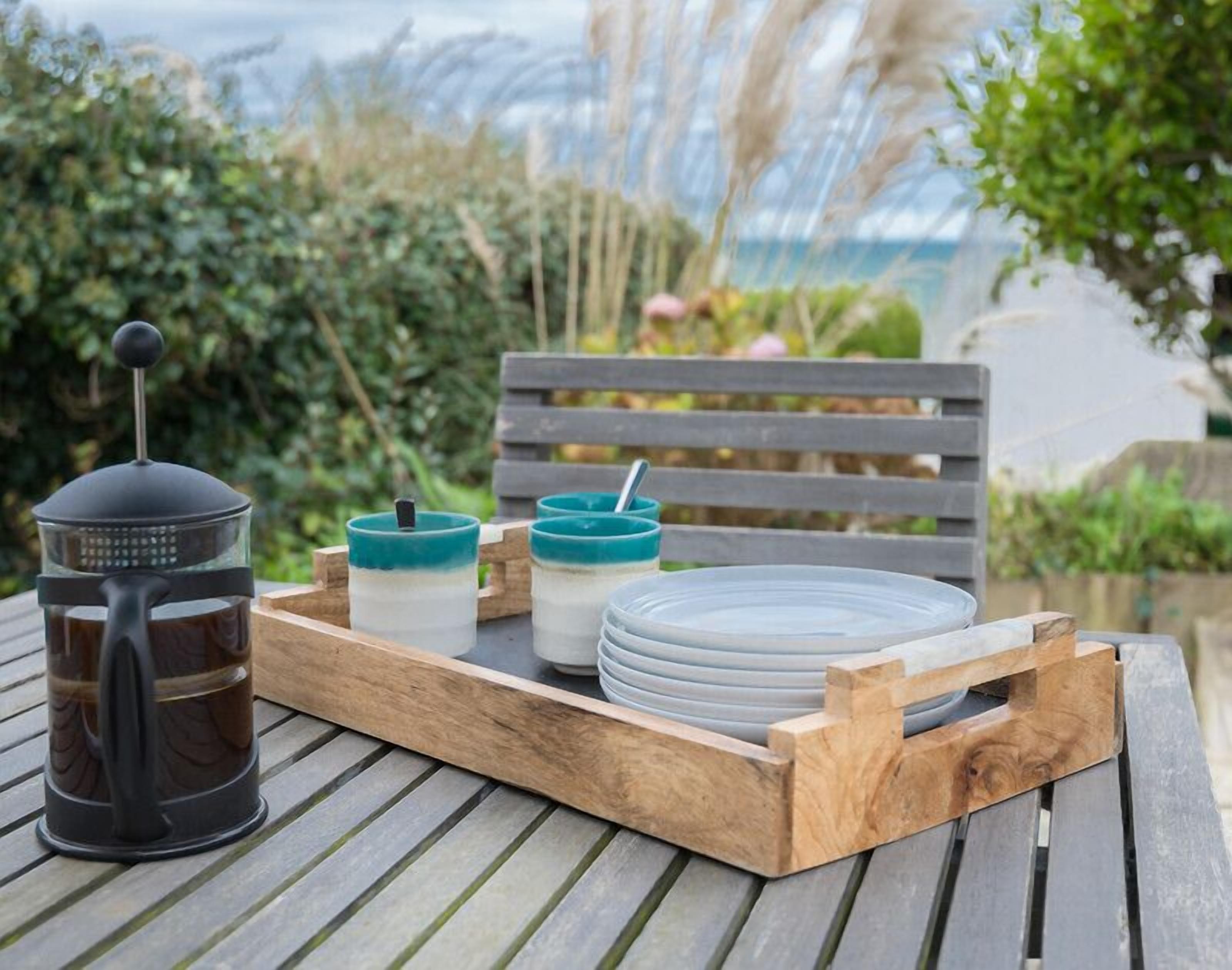 A wooden tray with a French press, two glass containers, and plates on a table outdoors.