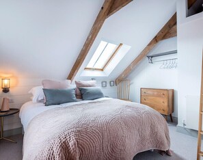 A bedroom with a sloped ceiling, a skylight, a bed with pillows, a bedside table with a lamp, and a wooden dresser. - Albany (Port Isaac)