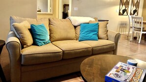 Un salon avec un canapé d’angle beige, deux coussins bleus, une table basse et un jeu de société.