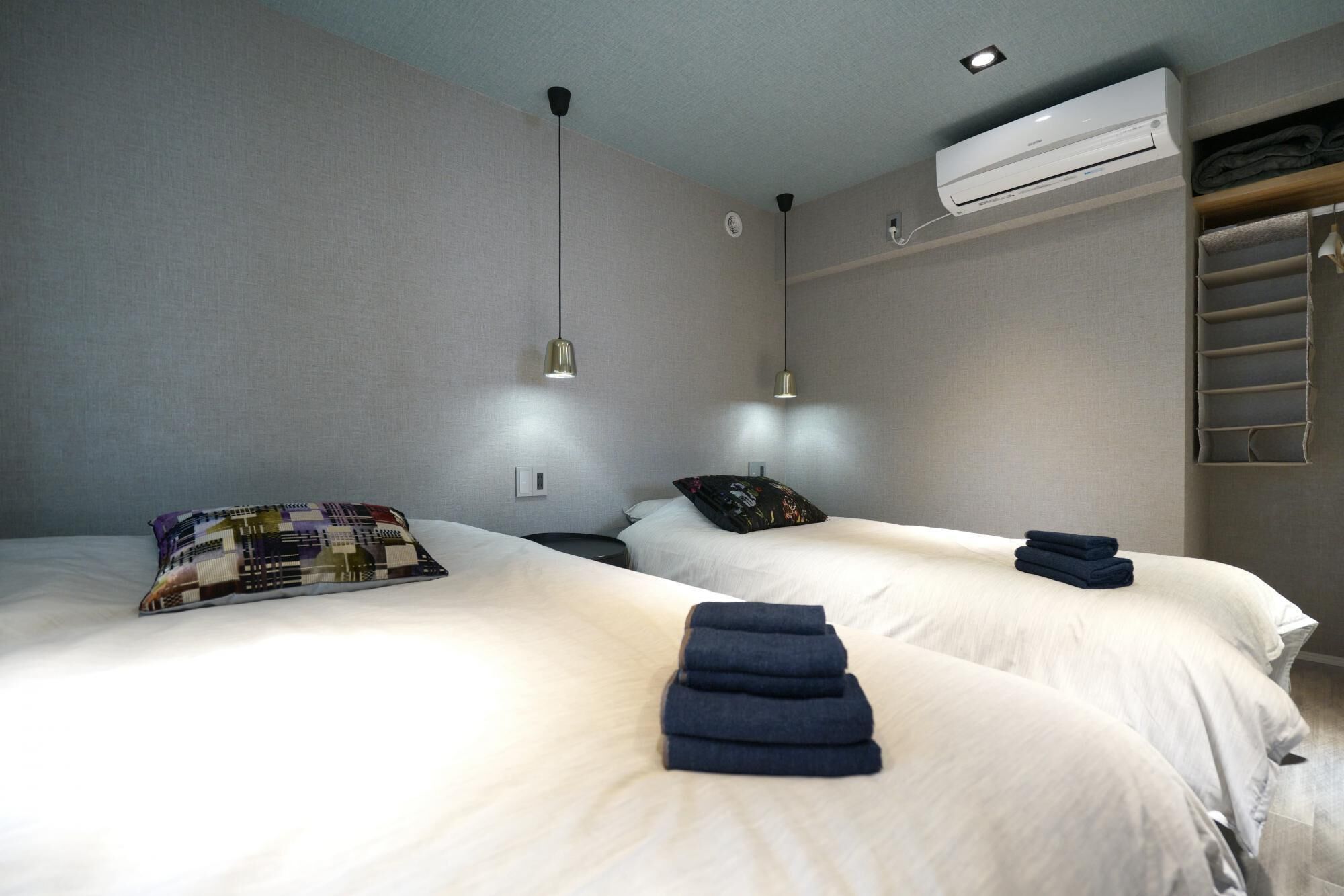 A hotel room with two beds, a ceiling fan, an air conditioning unit, and a built-in shelf with towels.