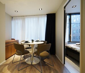 Quarto pequeno e moderno com uma mesa redonda e quatro cadeiras, janela com cortinas, cama com vista para edifícios e um armário de madeira.
