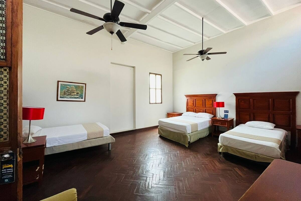 Ein Hotelzimmer mit zwei Einzelbetten, einem Holzkopfstück, einem Schreibtisch aus Holz, einer roten Lampe und einem Deckenventilator.