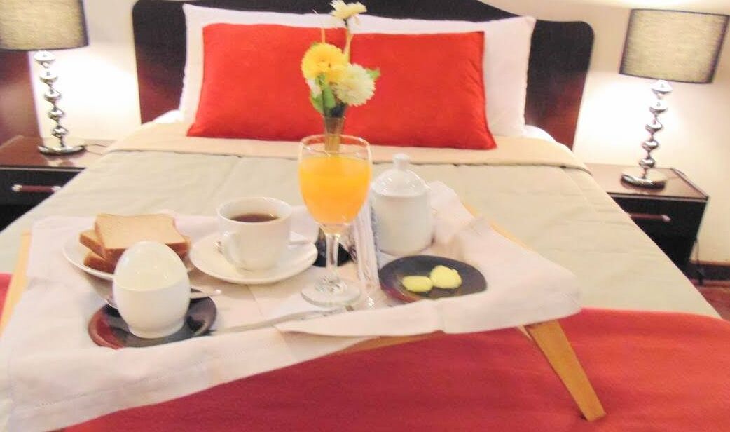 A neatly arranged breakfast tray on a bed with a red pillow, a glass of orange juice, toast, a cup of coffee, and a small plate with butter.