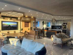 A modern living room with a dining area, kitchen, and a large flat-screen TV mounted on the wall. - SUNRISE with large terrace and far-reaching views (Pirmasens-Erlenbrunn)