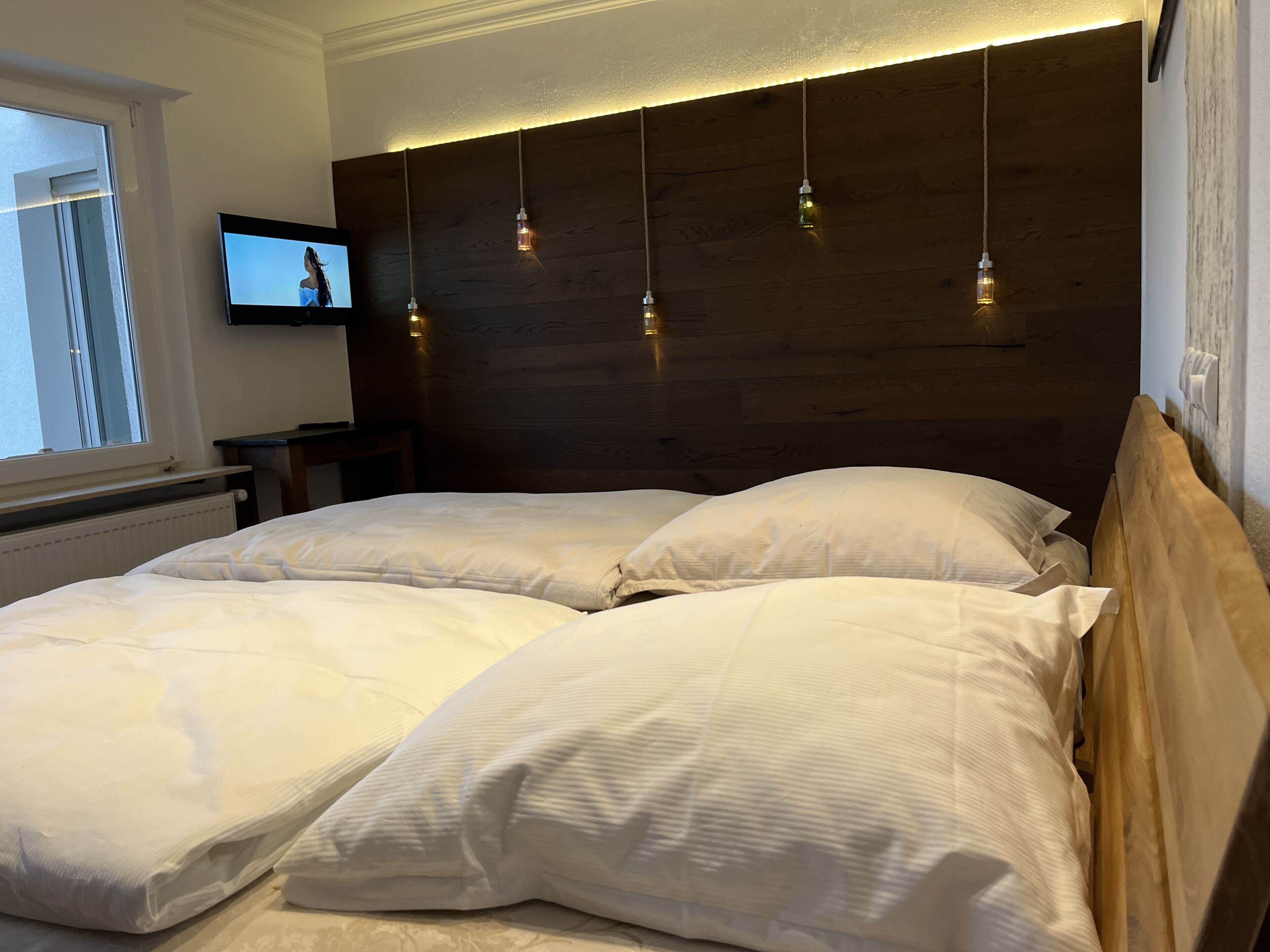 Une chambre à coucher avec une tête de lit en bois, un téléviseur à écran plat fixé au mur et une fenêtre avec des stores.
