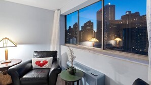 Een moderne hotelkamer met een groot raam met uitzicht op de stad, een leren bank met een hartvormig kussen, een kleine tafel met een plant en een staande verwarming.
