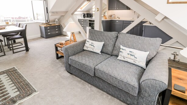 A loft-style living area with a gray sofa, a dining table, and a kitchen in the background. - The Sail Loft (Porthmadog)