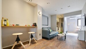 A modern living room with a grey sofa, two stools, a coffee table, and a kitchen area with a fridge and cabinets. - Real Life Meguro River Superb 2BR 6pax Shibuya (Tokyo)