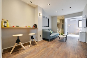 A modern living room with a grey sofa, two stools, a coffee table, and a kitchen area with a fridge and cabinets.