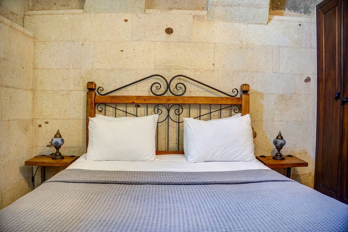 Una cama con cabecera de madera, dos almohadas blancas y una colcha estampada. Hay dos mesitas de noche con lámparas decorativas.