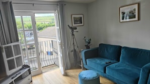 A living room with a blue sofa, a television, and a balcony with a view of greenery. - Castleview Large 3 Bedroom Family House - Glenarm (Ballymena)