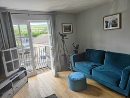 Una sala de estar con un sofá azul, un televisor y un balcón con vistas a zonas verdes.