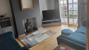 A living room with a fireplace, a TV, a sofa, and a large window with a view of buildings and greenery. - Castleview Large 3 Bedroom Family House - Glenarm (Ballymena)