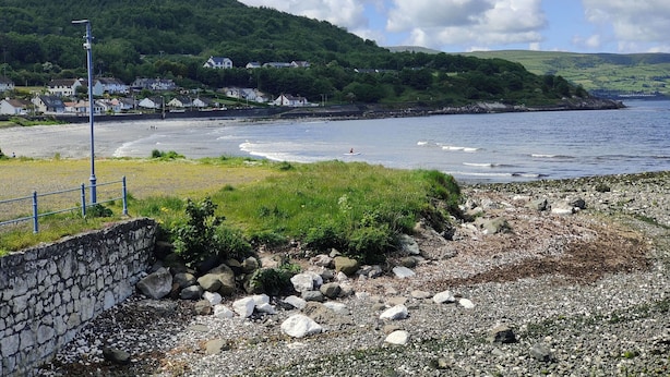 A coastal landscape with a rocky beach, a stone wall, and houses on a hillside. - Castleview Large 3 Bedroom Family House - Glenarm (Ballymena)