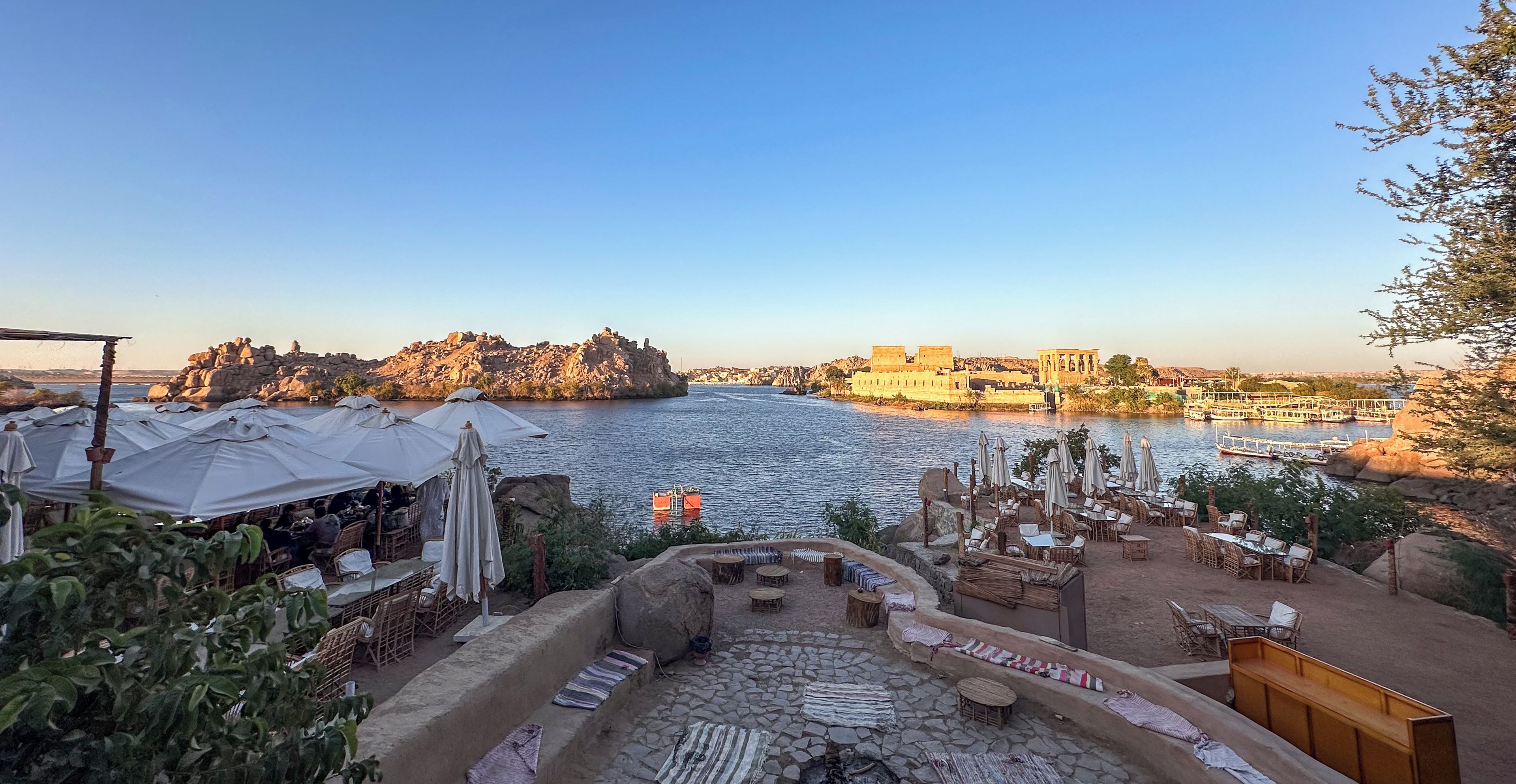 Une terrasse au bord de la rivière, avec des places assises en plein air et une vue sur un paysage urbain historique.