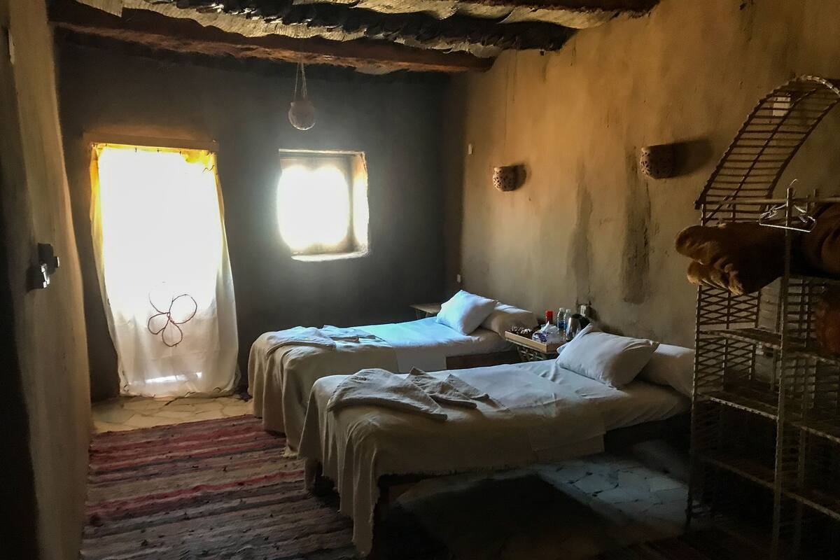 A room with two beds, a window, a hanging light, and a metal shelf with items.