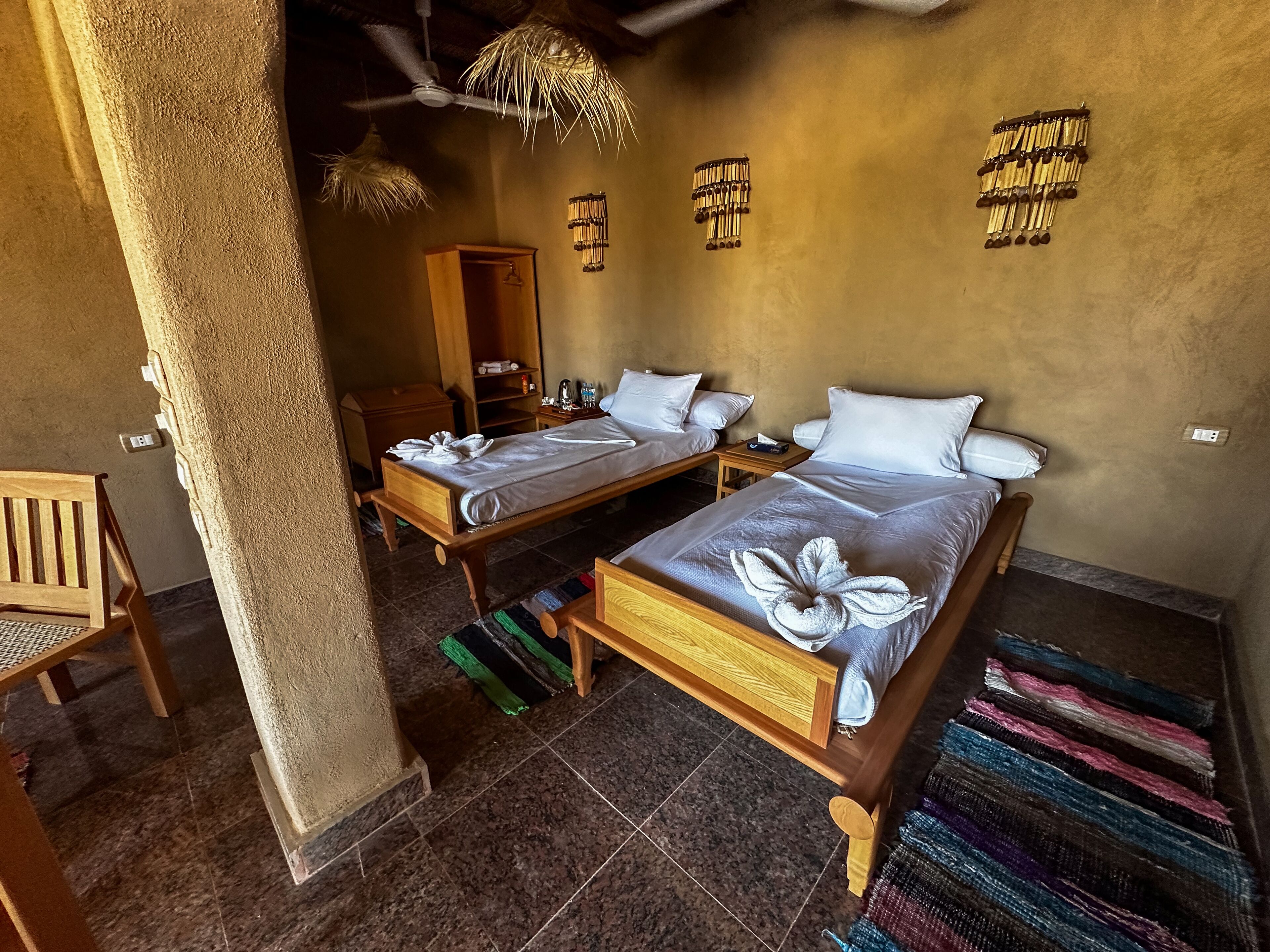 Ein Hotelzimmer mit zwei Holzbetten, einem Holzstuhl, einem kleinen Tisch und einem Deckenventilator.