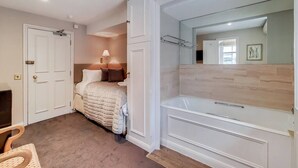 Ein Schlafzimmer mit einem Bett, einem Nachttisch, einer Lampe und einem großen Spiegel über einer Badewanne.