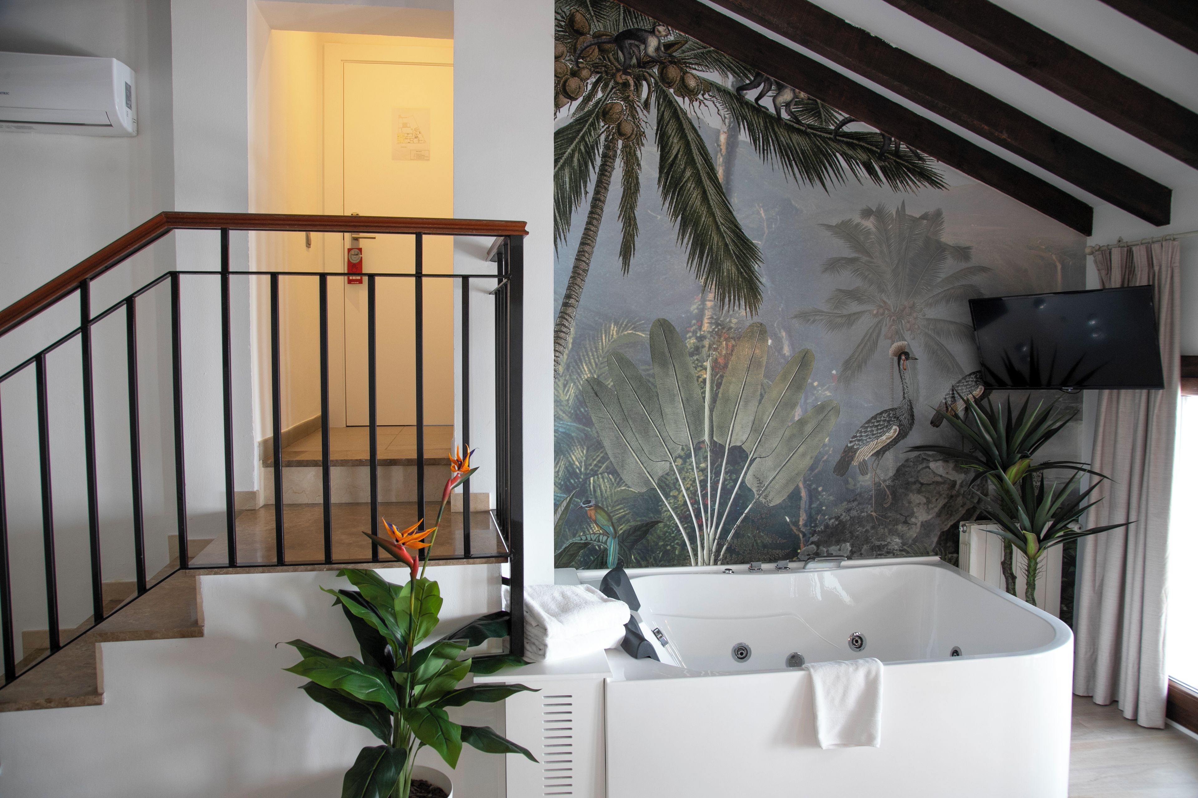 A modern bathroom with a large bathtub, a staircase with a wooden railing, and a tropical mural on the wall.