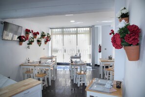 Un comedor con mesas y sillas blancas, un ventanal con persianas y plantas en macetas en las paredes.