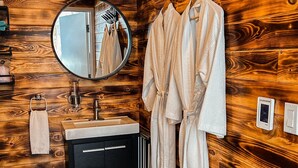 A bathroom with wooden walls, a round mirror, a sink, and two white bathrobes hanging on a rack. - Eagles Nest Converted Shipping Container at Sally's Brook Wilderness Cabins (Englishtown)