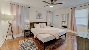 Quarto com cama, mesas de cabeceira, luminária de chão, ventilador de teto e janela com cortinas.