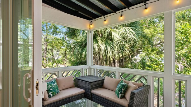 A covered outdoor seating area with wicker furniture and glass tables, surrounded by white railings and large windows offering views of greenery. - Zen-N-Sea offers poolside relaxation and near the beach entrance, sleeps 8 (St Simons Island)