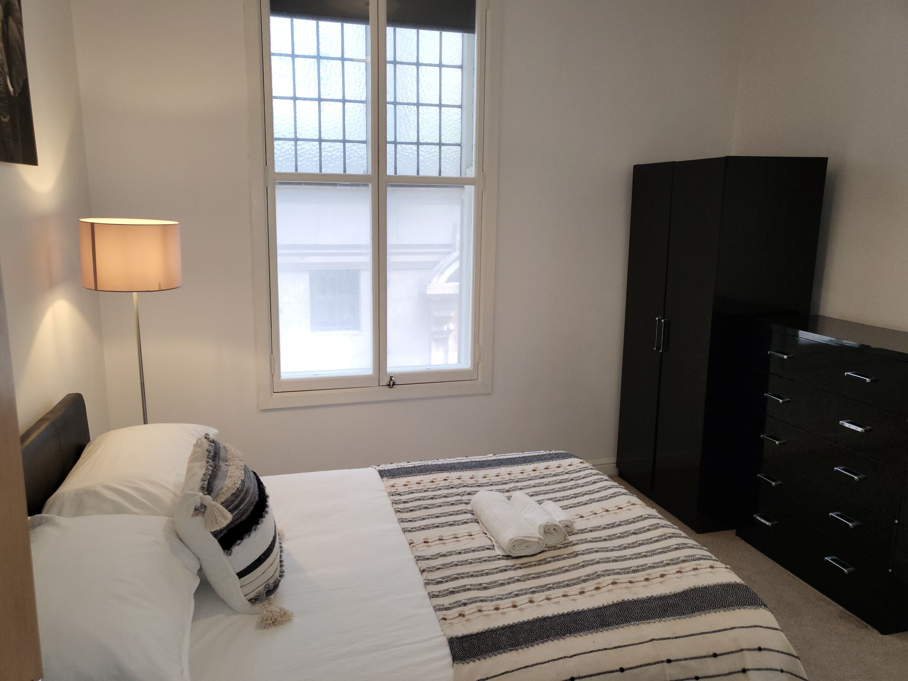 A bedroom with a bed, a nightstand, a lamp, a window, and a wardrobe.