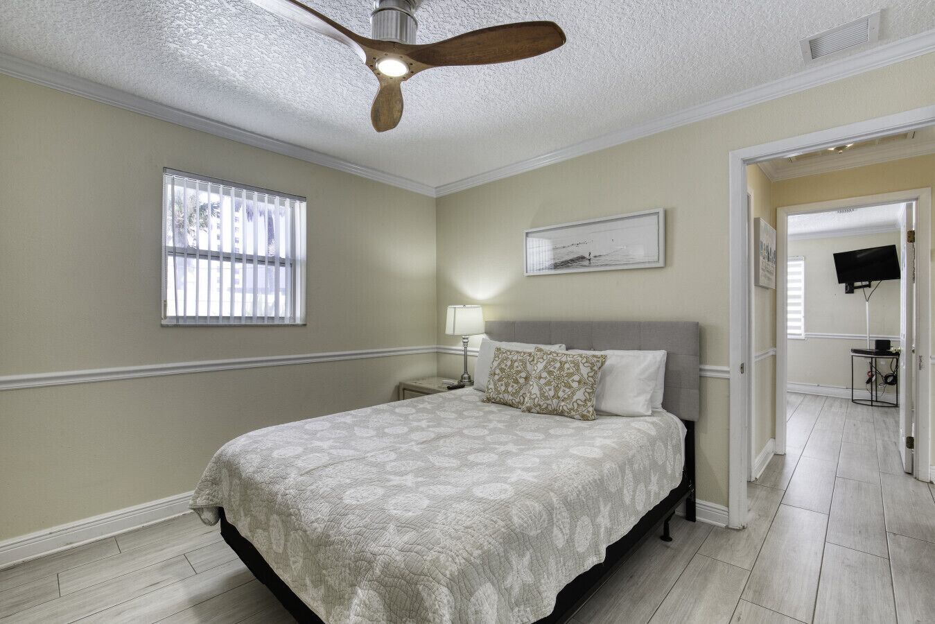 A bedroom with a bed, a ceiling fan, a window with blinds, a bedside table with a lamp, and a television in the adjacent room.