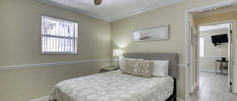 A bedroom with a bed, a ceiling fan, a window with blinds, a bedside table with a lamp, and a television in the adjacent room.