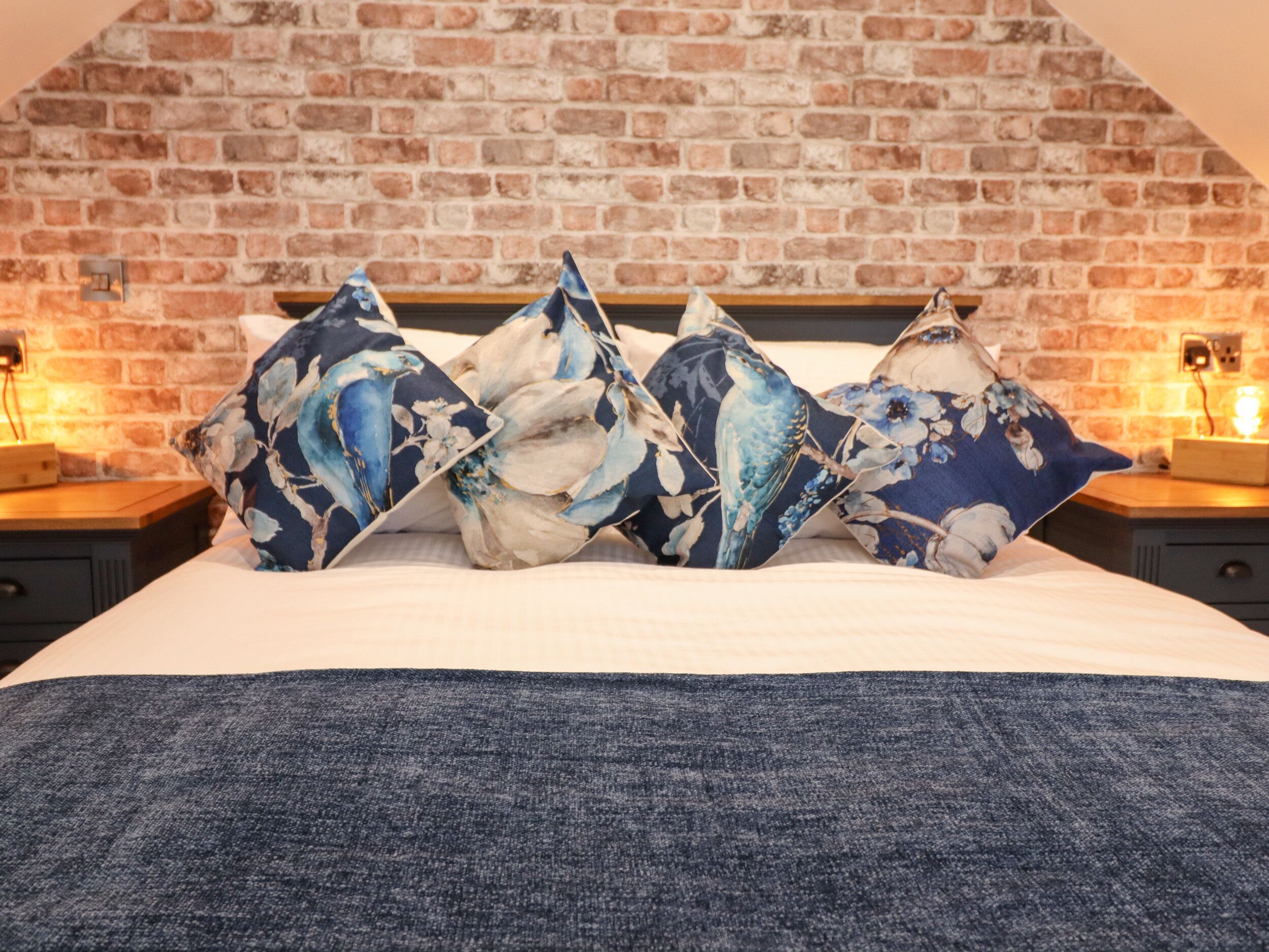 A bed with floral-patterned pillows, a brick wall, and a wooden headboard.