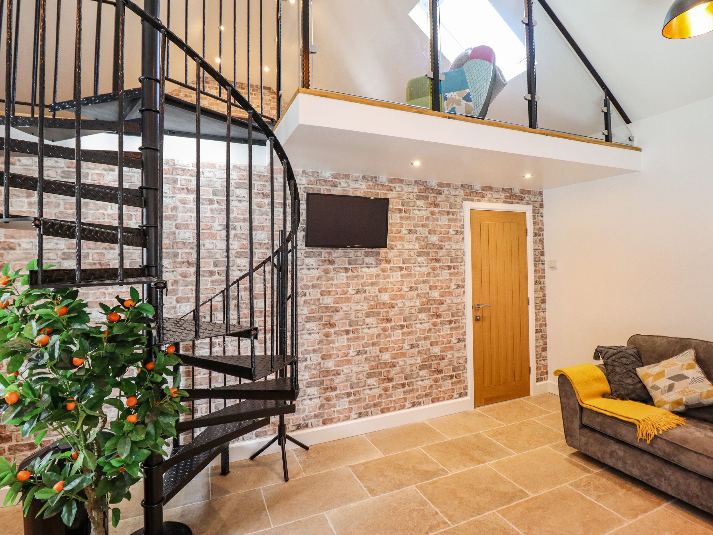 A spacious living area with a brick wall, a wooden door, a black spiral staircase, and a sofa with a yellow throw blanket.