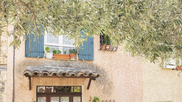 A traditional building with a wooden door, a window with patterned curtains, and potted plants. - House l'oustaou les 2 oliviers (Moustiers-Sainte-Marie)