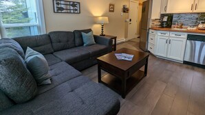 Una sala de estar con un sofá modular, una mesa de centro y una cocina con armarios blancos y un refrigerador.