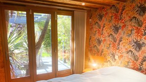 A hotel room with a bed, a window with a view of greenery, and a tropical wallpaper. - Zen Garden Cottage - Sleeps 2 - Patio & Parking (Poole)
