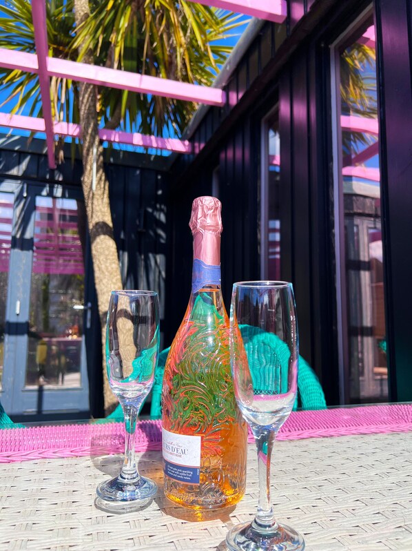 A bottle of fruit brandy and two glasses on a table with a woven mat, under a pink pergola with palm trees in the background.