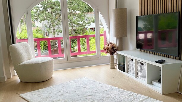 A modern living room with a large arched window, a white sofa, a white rug, a TV on a white stand, and a wooden wall panel. - 4 bedroom villa in la baule-escoublac (La Baule-Escoublac)