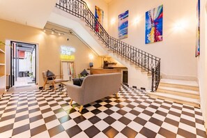 Eine Hotelhalle mit Schachbrettfußboden, einem grauen Sofa, einer Treppe mit schwarzen Geländern und farbenfrohen abstrakten Gemälden an den Wänden.