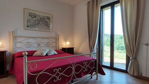 Un dormitorio con estructura de cama metálica, colcha roja, dos mesitas de noche con lámparas, una ventana con cortinas, y un cuadro enmarcado en la pared.