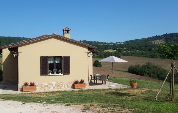A small house with a tiled roof, a chimney, and a window with shutters. There is a patio with a table and chairs, and a large umbrella. The house is surrounded by a grassy area and a hilly landscape in the background.