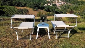 Tres tumbonas blancas con una mesa y una botella de vino en el centro, con vistas a un viñedo y un pueblo en la colina.