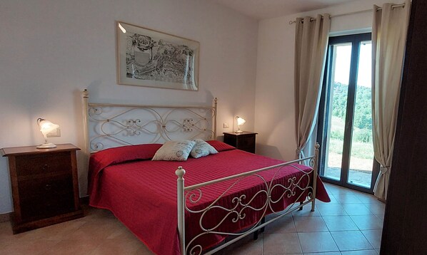 A bedroom with a metal bed frame, red bedding, two bedside lamps, a nightstand, a framed picture on the wall, and a view through a window with curtains.