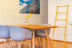 Een eetgedeelte met een houten tafel, twee grijze stoelen, een vaas met gele bloemen en een gele ladder tegen een muur met een stadsgezicht schilderij.