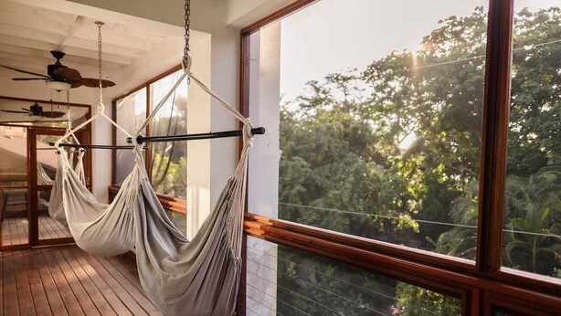 Una hamaca colgada de cadenas en el techo en una habitación con suelo de madera y grandes ventanales con vistas a un bosque.